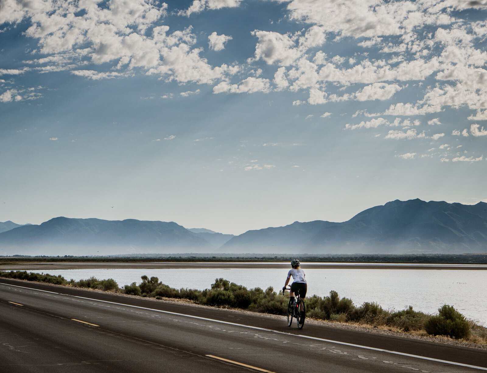 A cyclist on a lakeside road beneath mountains