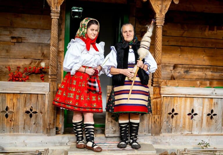 Two women in traditional Romanian folk costume on a wooden porch